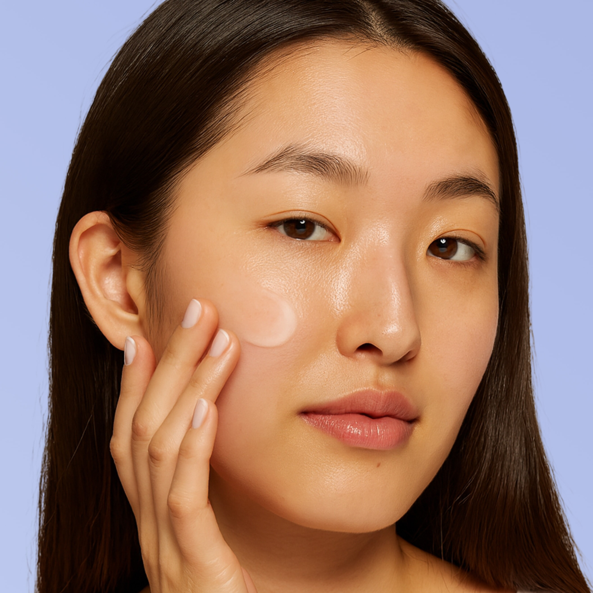 Close-up of a woman with smooth, glowing skin gently applying a small amount of cream to her cheek, demonstrating a hydrating skincare step against a soft lavender background.