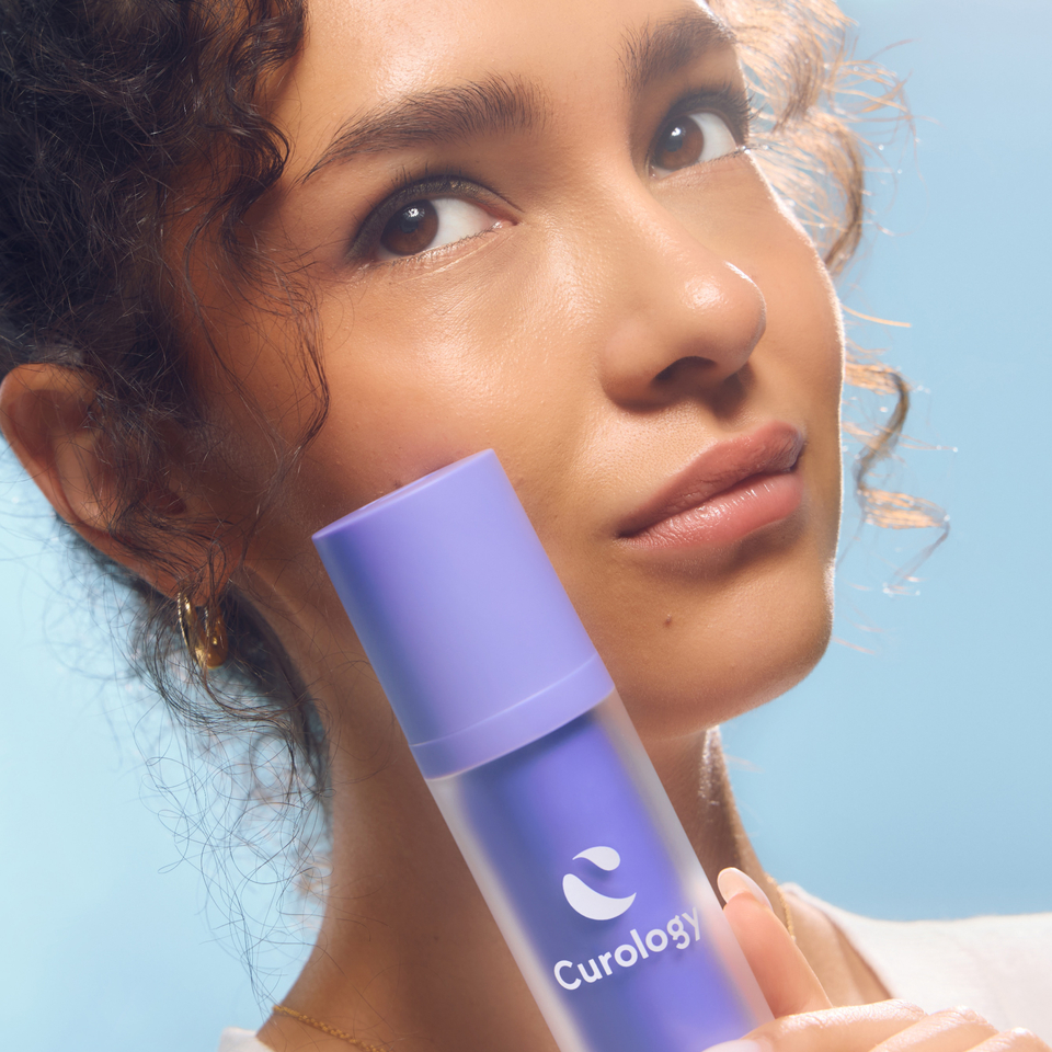 A young woman holds a purple cylindrical bottle of Curology DayPrime Rx